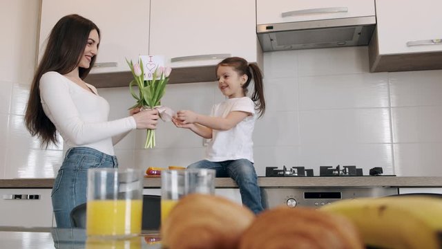 Little girl has a present for her mother. She is giving the mother a bunch of flowers and greet her with the day of mother. The mother is surprised and happy.