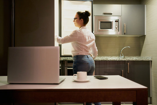 A young woman who works with a laptop looks in the fridge