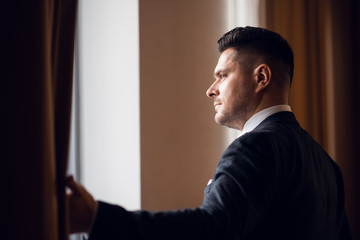 A young businessman in a suit parting the curtains in his hotel room to let the morning light in