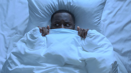 Adult african american man scared of noise hiding under blanket, infantility