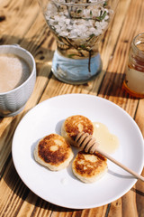 Delicious three cottage cheese pancakes with honey lying on the white plate with the branch of a flowering tree on the wooden table background with the cuo of coffee. Curd cheese fritters.
