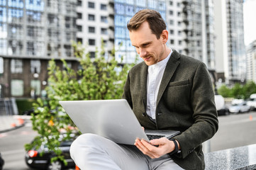 Work outdoors. A young man in smart casual clothes uses a laptop for work, a skyscraper in the background