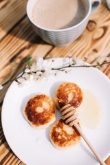 Delicious three cottage cheese pancakes with honey lying on the white plate with the branch of a flowering tree on the wooden table background with the cuo of coffee. Curd cheese fritters.