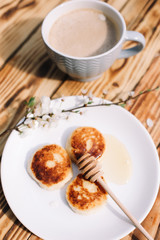 Delicious three cottage cheese pancakes with honey lying on the white plate with the branch of a flowering tree on the wooden table background with the cuo of coffee. Curd cheese fritters.