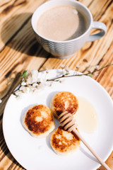 Delicious three cottage cheese pancakes with honey lying on the white plate with the branch of a flowering tree on the wooden table background with the cuo of coffee. Curd cheese fritters.