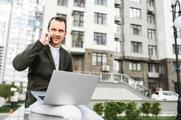 Young positive businessman uses a laptop to work outdoors, he sits and speaks on the phone with a the cityscape on background