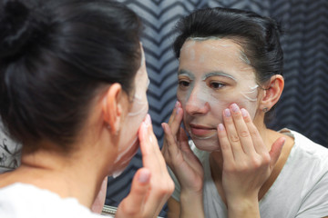 A girl in a cosmetic mask looks in the mirror at her reflection. Makes home beauty treatments