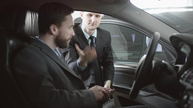 Handsome man is sitting in a car, talking to the car dealer. Trader gives key, shake shands with auto dealership customer Young car dealer serving customer in showroom.