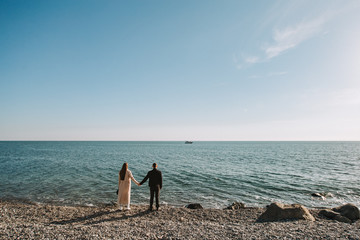 A full-length couple in love stands on the seashore, holding hands and looking into the distance at a floating ship. The couple stands on a warm Sunny day against a background of blue sky and calm sea © Ярослав Яровой