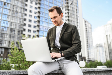 Work outdoors. A young man in smart casual clothes uses a laptop for work, a skyscraper in the background