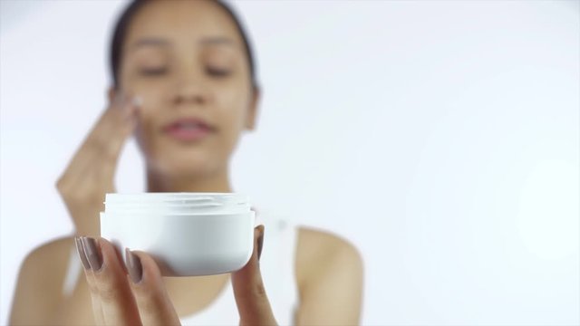 Closeup shot of a happy girl applying moisturizer on her face with a jar in focus - Skin Care. Beautiful Indian female rubbing cream on her cheeks while holding a jar in her hand - white background...