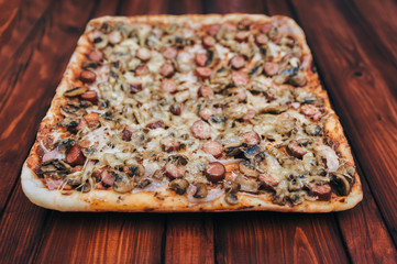 Homemade rectangular pizza with sausages, mushrooms, cheese and onions lies on a mahogany wooden table. The concept of lunch, fast food and Italian cuisine.