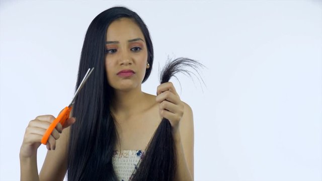 Young Indian girl tired of hair damage thinking of cutting split ends - haircare concept. Frustrated woman with hair problems planning to trim her hair while holding the scissors in her hand - whit...