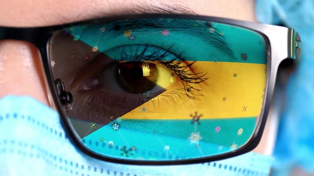 closeup, eye, part of doctor face in medical mask, glasses, which painted in colors of Bahamas flag. Many viruses, germs moving on glass.State interests in vaccines, drugs invention, pathogenic