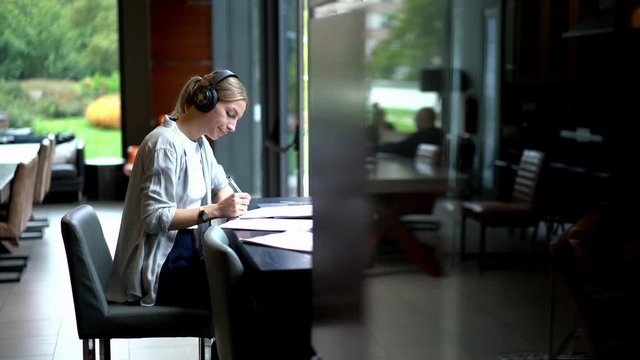 Intelligent millennial woman sitting at comfortable desktop and writing information for university course work, smart female student in electronic headphones for noise cancellation listening audio
