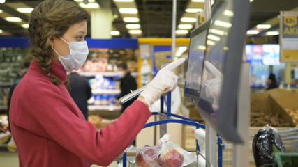Woman in a medical mask and rubber gloves weighs apples on an electronic scale in a supermarket. Coronavirus pandemic protection. In the background are buyers in protective masks.