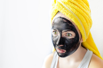 Surprised teen girl portrait with black facial clay mask and yellow towel on head over white background. Spa beauty treatments at home, skin care, positive emotion, facial expression. Copy space.