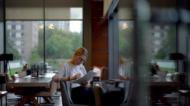 Slow motion effect of intelligent woman in classic spectacles reading information from univeristy report and editing text during learning time, concept of knowledge and education
