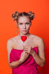 The girl in a red dress on a orange background in the studio. Blonde girl with two hair knots holding red heart on the stick.