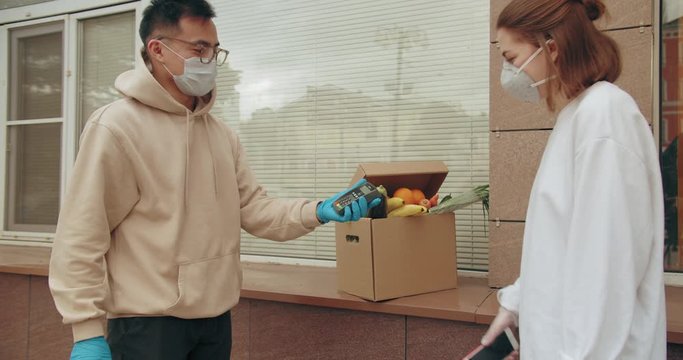 postman or delivery asian man carry small box deliver to young woman customer at home contactless nfc terminal payment. Man wearing mask prevent covid or coranavirus quarantine pandemic. 