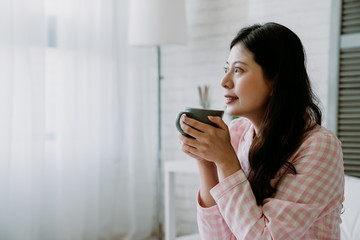 pretty asian lady holding coffee with both hands is enjoying her hot drink and the refreshing air in early morning. korean lady in plaid pajamas looking outside window with a smile.