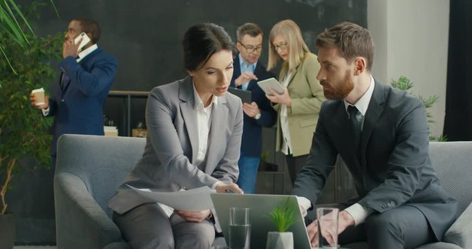 Young businessman and businesswoman sitting at hall in break of conference at laptop computer and talking over partnership. Caucasian male and female business partners brainstorming on strategy.