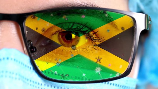 closeup, eye, part of doctor face in medical mask, glasses, which painted in colors of Jamaica flag. Many viruses, germs moving on glass.State interests in vaccines, drugs invention, pathogenic