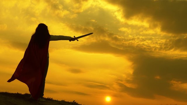 free woman knight. super woman with sword in his hand and in a red cloak stands on mountain in sunset light. free woman playing superhero. girl plays roman lenin in bright rays of sun against sky
