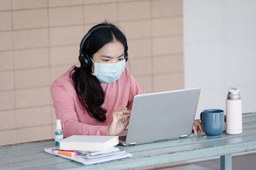 Portrait of a young Asian female adult learner studying online at home during Coronavirus or COVID-19  pandemic. Businesswoman wear mask works with laptop at home in house corridor and garden.