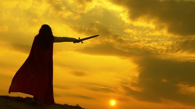 free woman knight. super woman with sword in his hand and in a red cloak stands on mountain in sunset light. free woman playing superhero. girl plays roman lenin in bright rays of sun against sky