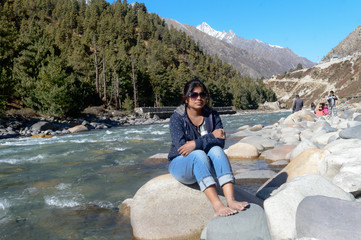 Solo Indian traveler in winter casual sitting alone near stream flowing water mountain valley. Snow capped Himalayan mountain forest blue sky in background. Spiti Valley Himachal Pradesh India Asia