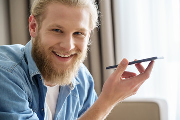 Happy young man holding smart phone using virtual digital voice assistant look at camera at home....