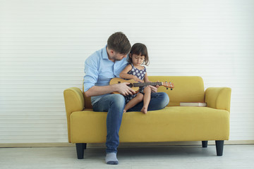 Obraz premium Young handsome Father is teaching his little cute daughter to playing ukulele guitar on the yellow sofa at home