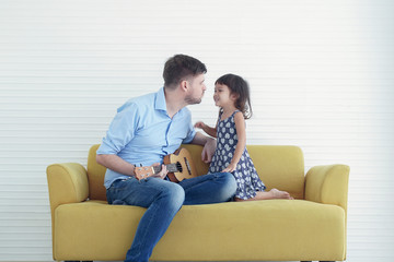 Little cute daughter looking at her Young handsome father while he is playing ukulele guitar together on the yellow sofa at home
