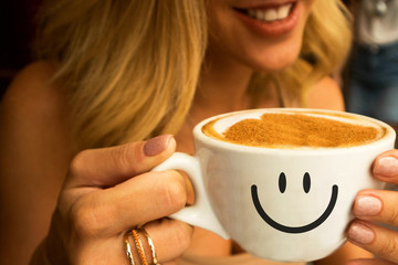 smiling young girl in a cafe  holding a big cup of hot cappuccino with coffee foam design of heart on it and smile on the cup
