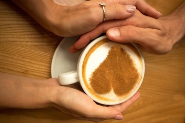 romantic lovely young couple in love sit in cafe having breakfast,closeup hands holding a cup of coffee with art design heart on it   on a wooden table background 