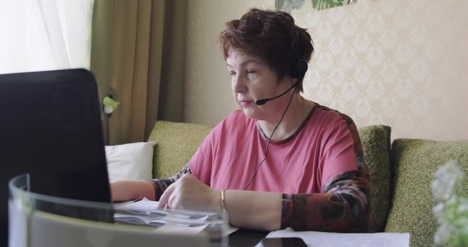 Modern grandmother uses laptop, Internet, headset to communicate on Internet, modern person, home office, remote work, pensioner knows how to manage new technologies, advanced user, videoconference