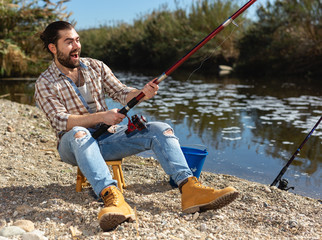 Positive man sitting near river and holding fishing rod