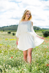 Fototapeta na wymiar Pretty girl wearing white dress walking in field of wild camomile flowers