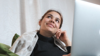 Cute attractive girl dreaming and fantasizing while using a computer at home. Smiling woman working from her couch and feeling cozy. 