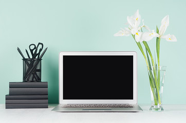Fresh spring workplace for work and study with blank laptop display, black stationery, books and white bouquet in light green mint menthe interior on white wood desk.