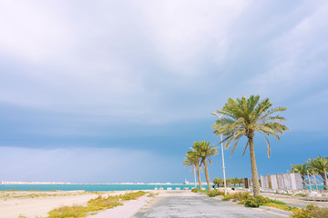 Street road to the sea beach with palm trees and electric poles on the side over clouds sky background, Bahrain.