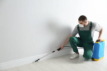 Pest control worker in uniform spraying pesticide indoors. Space for text - obrazy, fototapety, plakaty