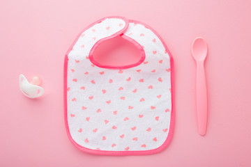 Baby textile bib, plastic spoon and soother on light pink table background. Pastel color. Closeup....