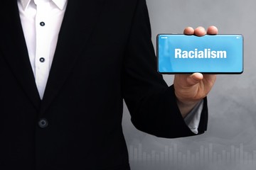 Racialism. Businessman in a suit holds a smartphone at the camera. The term Racialism is on the phone. Concept for business, finance, statistics, analysis, economy