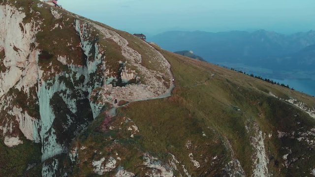 Orbital drone sunset clip at dramatic mountain peak with cross. Two hikers climbing at rocky cliff of Schafberg, Salzkammergut, Austria. Aerial 4K footage with Alpine view of Wolfgangsee lake