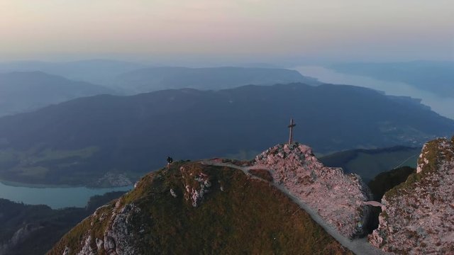 Orbital drone sunset shot at mountain peak and cross. Hiker girl climbing and taking pictures at rocky cliff Schafberg, Salzkammergut, Austria Aerial 4K footage dramatic Alpine view Attersee lake