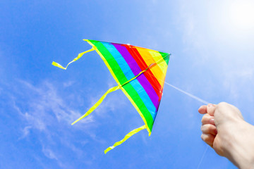 A hand is holding a rope, multi-colored stripes of a rainbow kite flying high in a clear blue sky...
