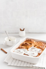 Fragrant freshly baked Cinnamon rolls or cinnabons in ceramic dish and white cream glaze in a cup on white background. Cinnabons recipe. Pastry, bakery. Side view, copy space