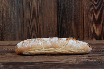 Crunchy, oblong, freshly baked bread sprinkled with flour, side view. A fresh, loaf of bread on brown, wooden, rustic boards.